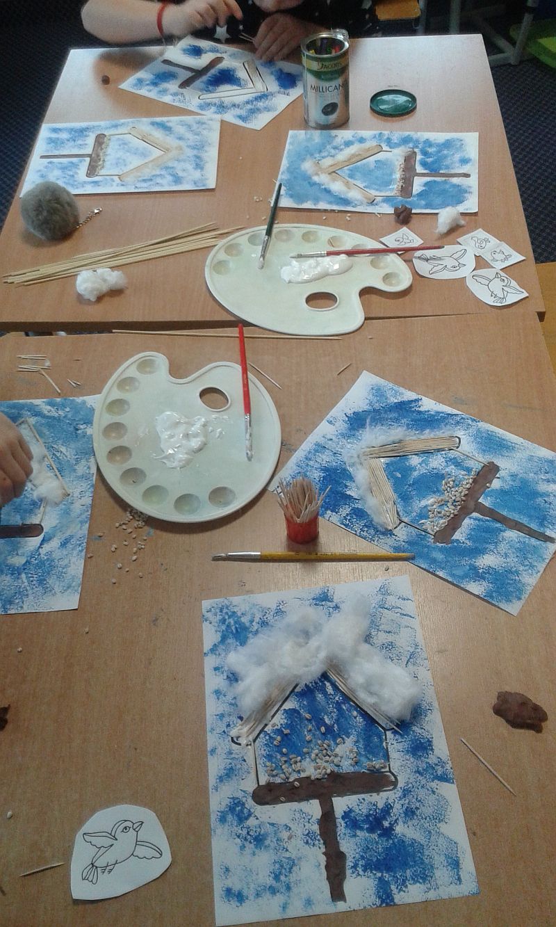 na ławkach są porozkładane prace dzieci, które dalej kończą. Na pracach na niebieskim tle są karmniki wykonane z wykałaczek, plasteliny, waty oraz kaszy 