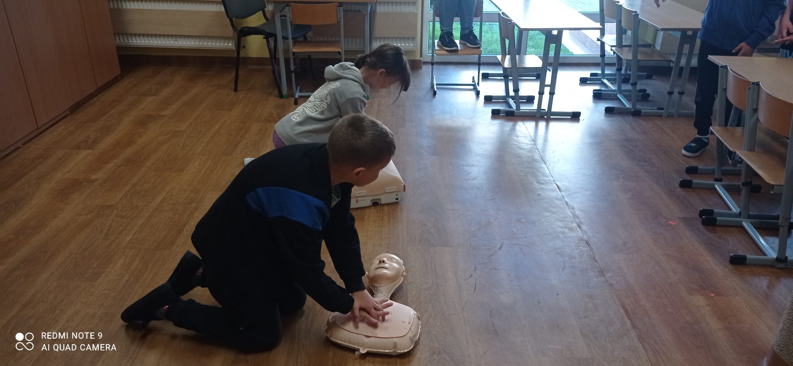 Zdjęcie przedstawia dwoje dzieci które klęcząc na podłodze ugniatają fantomy wykonując resuscytację