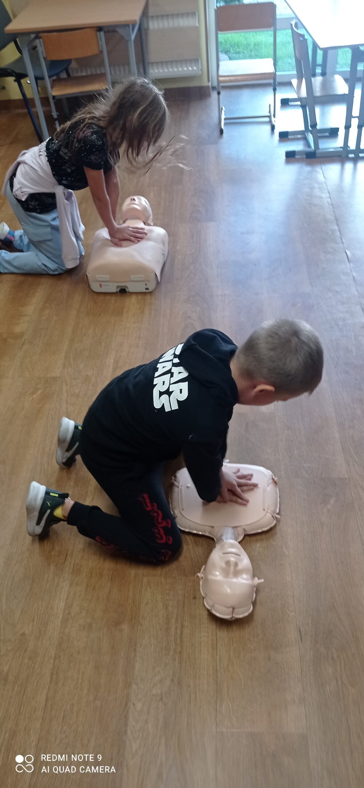 Zdjęcie przedstawia dwoje dzieci które klęcząc na podłodze ugniatają fantomy wykonując resuscytację