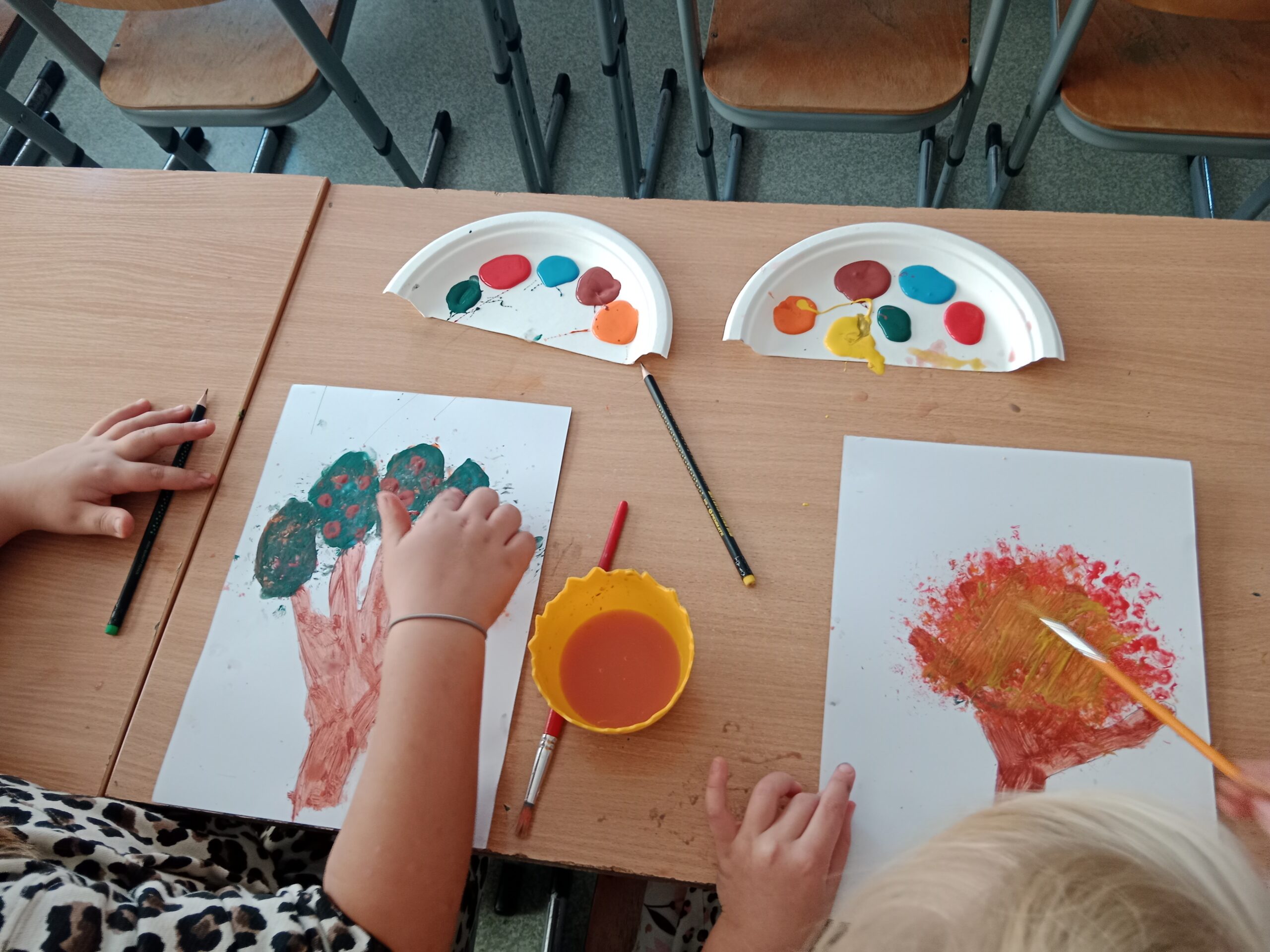 Zdjęcie przedstawia prace dzieci, która za pomocą odcisków palców maczanych w farbie wykonują na kartce papieru drzewka jesienne.