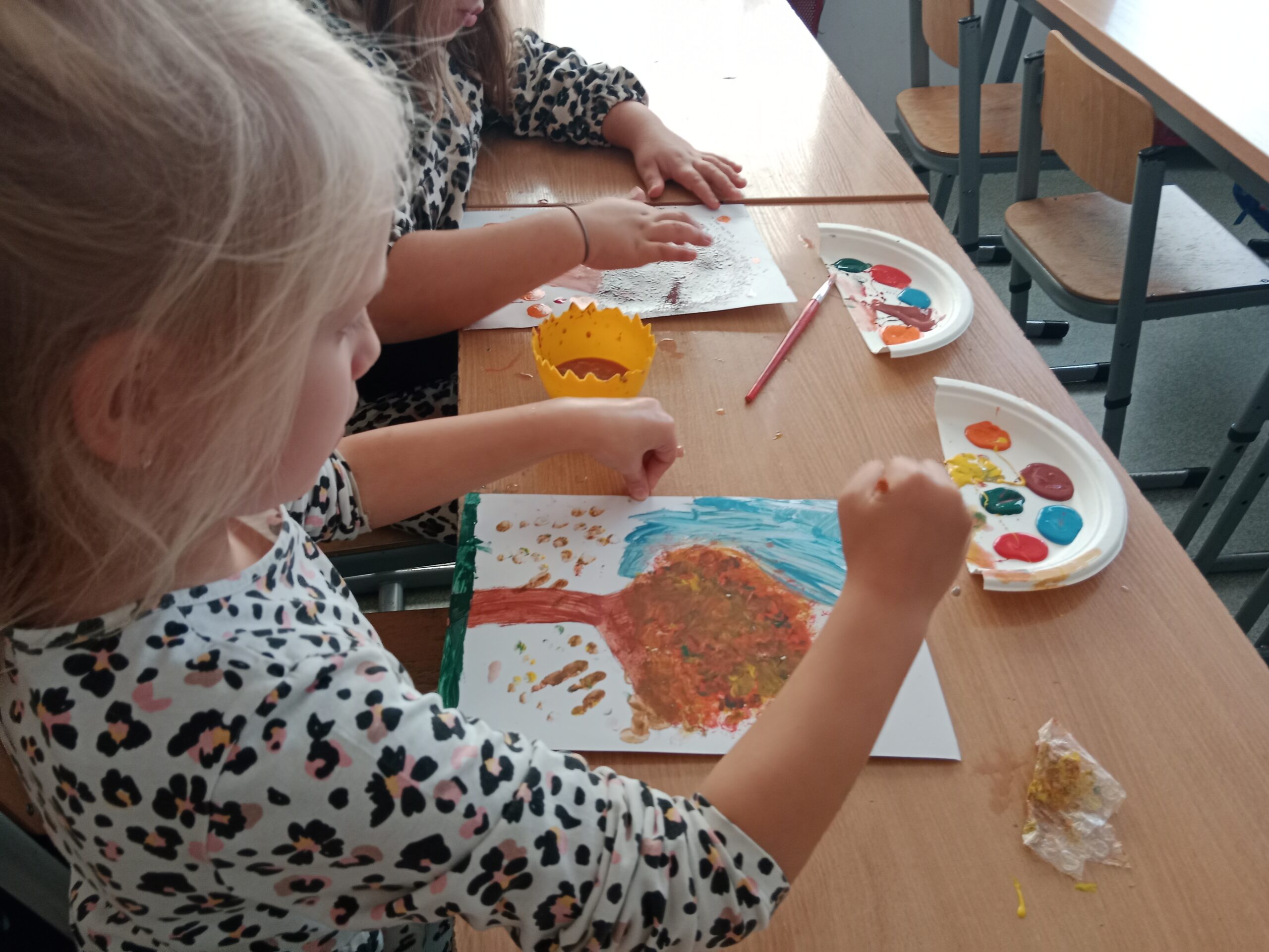 Zdjęcie przedstawia dziewczynkę, która za pomocą odcisków palców maczanych w farbie wykonuję na kartce papieru drzewko jesienne.