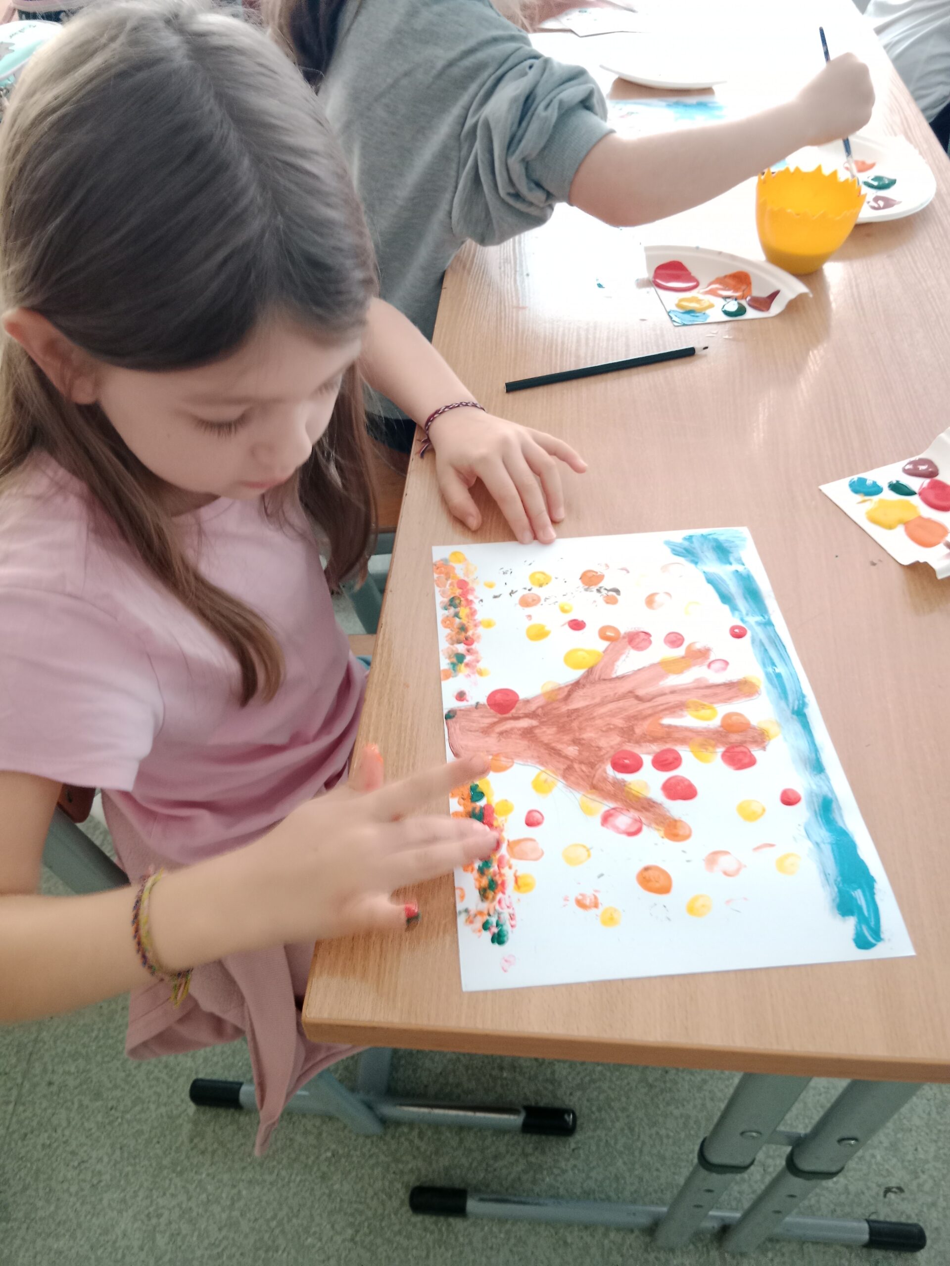 Zdjęcie przedstawia dziewczynkę, która za pomocą odcisków palców maczanych w farbie wykonuję na kartce papieru drzewko jesienne.