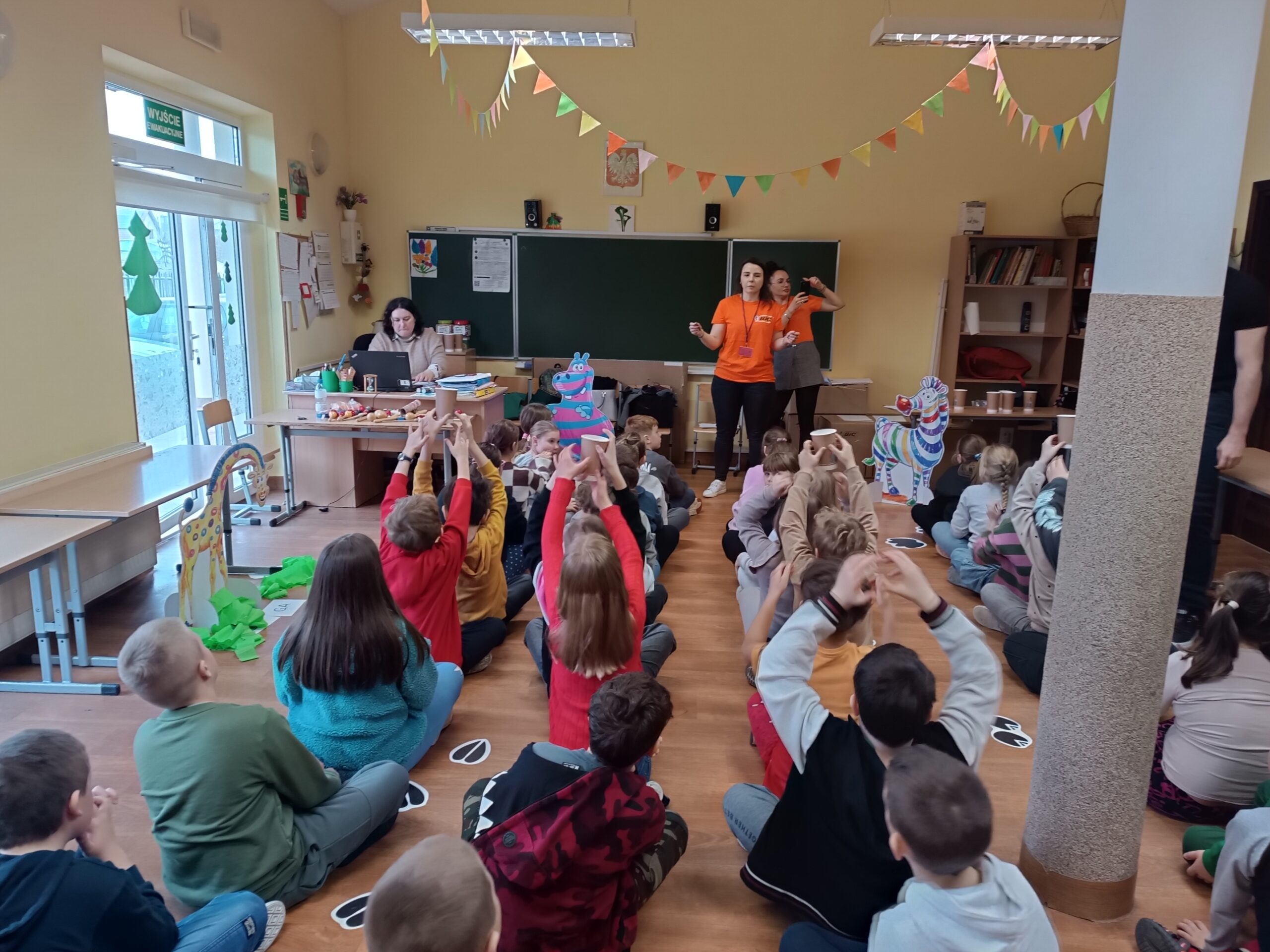 Na zdjęciu widać dzieci siedzące w rzędach, przekazujące sobie kubki z wodą nad głową 