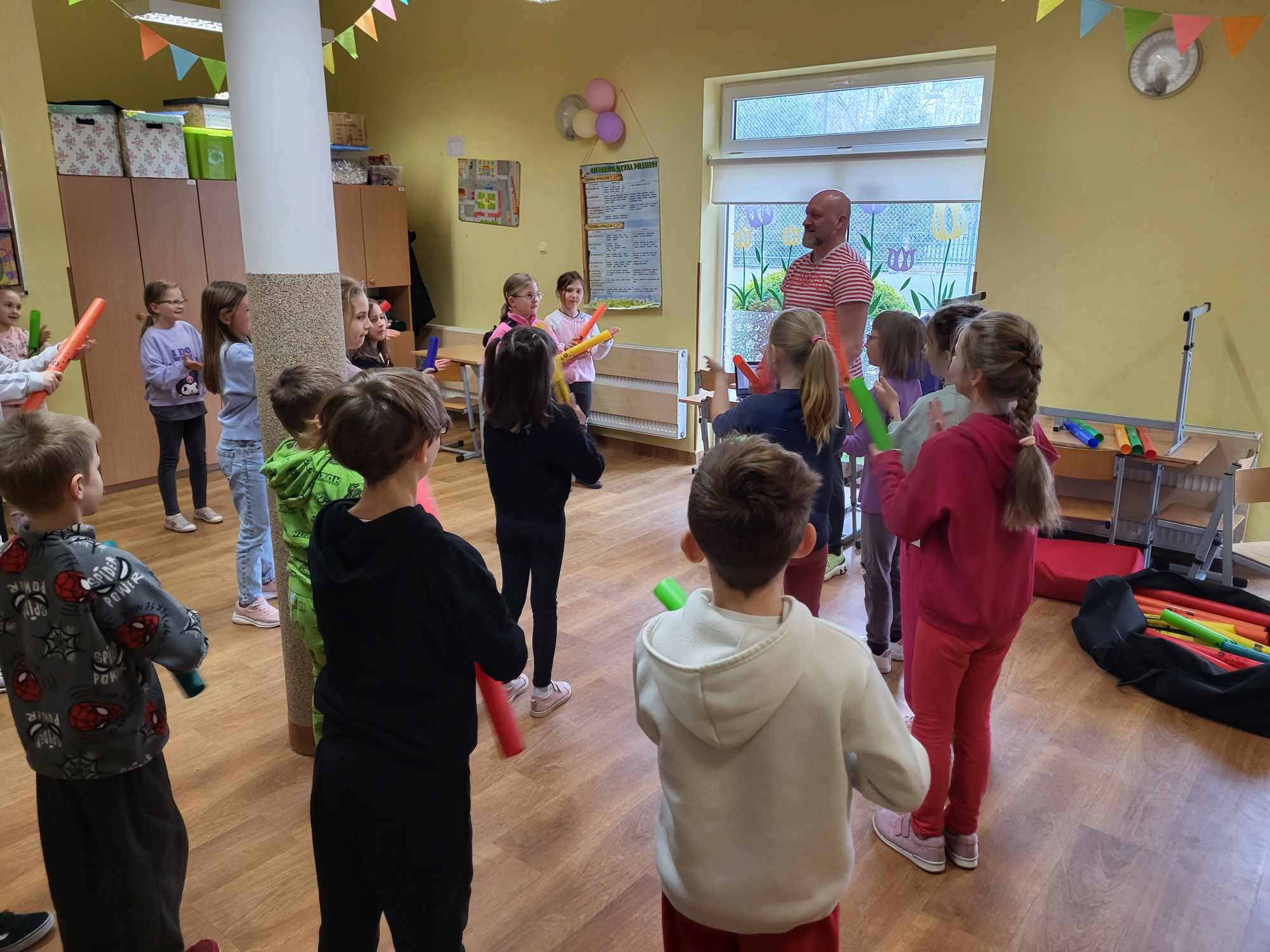 Na zdjęciu widać pana, który prowadzi warsztaty oraz grupę stojących dzieci, trzymających w rękach kolorowe rurki