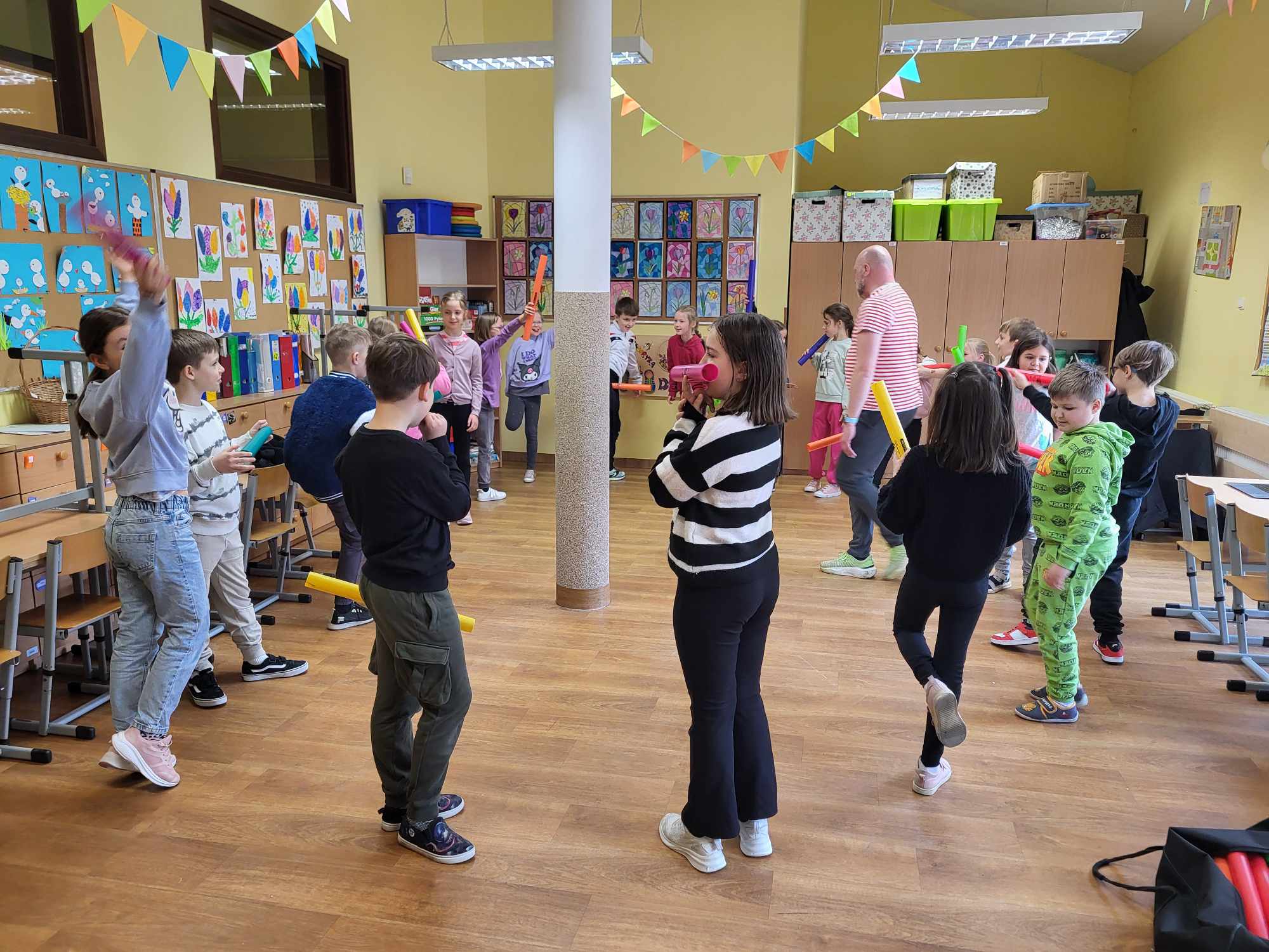 Na zdjęciu widać pana, który prowadzi warsztaty oraz grupę stojących dzieci, trzymających w rękach kolorowe rurki