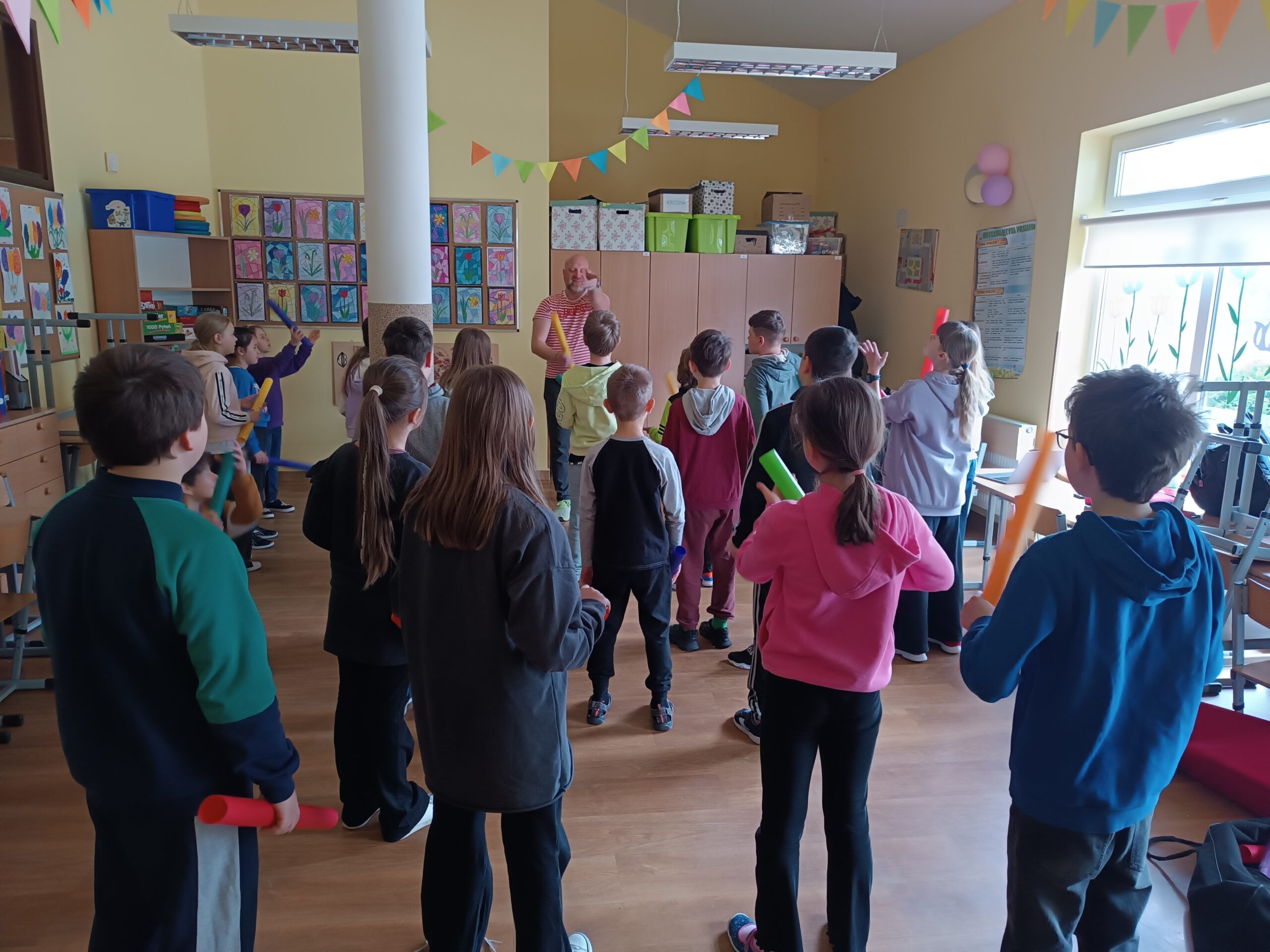 Na zdjęciu widać pana, który prowadzi warsztaty oraz grupę stojących dzieci, trzymających w rękach kolorowe rurki