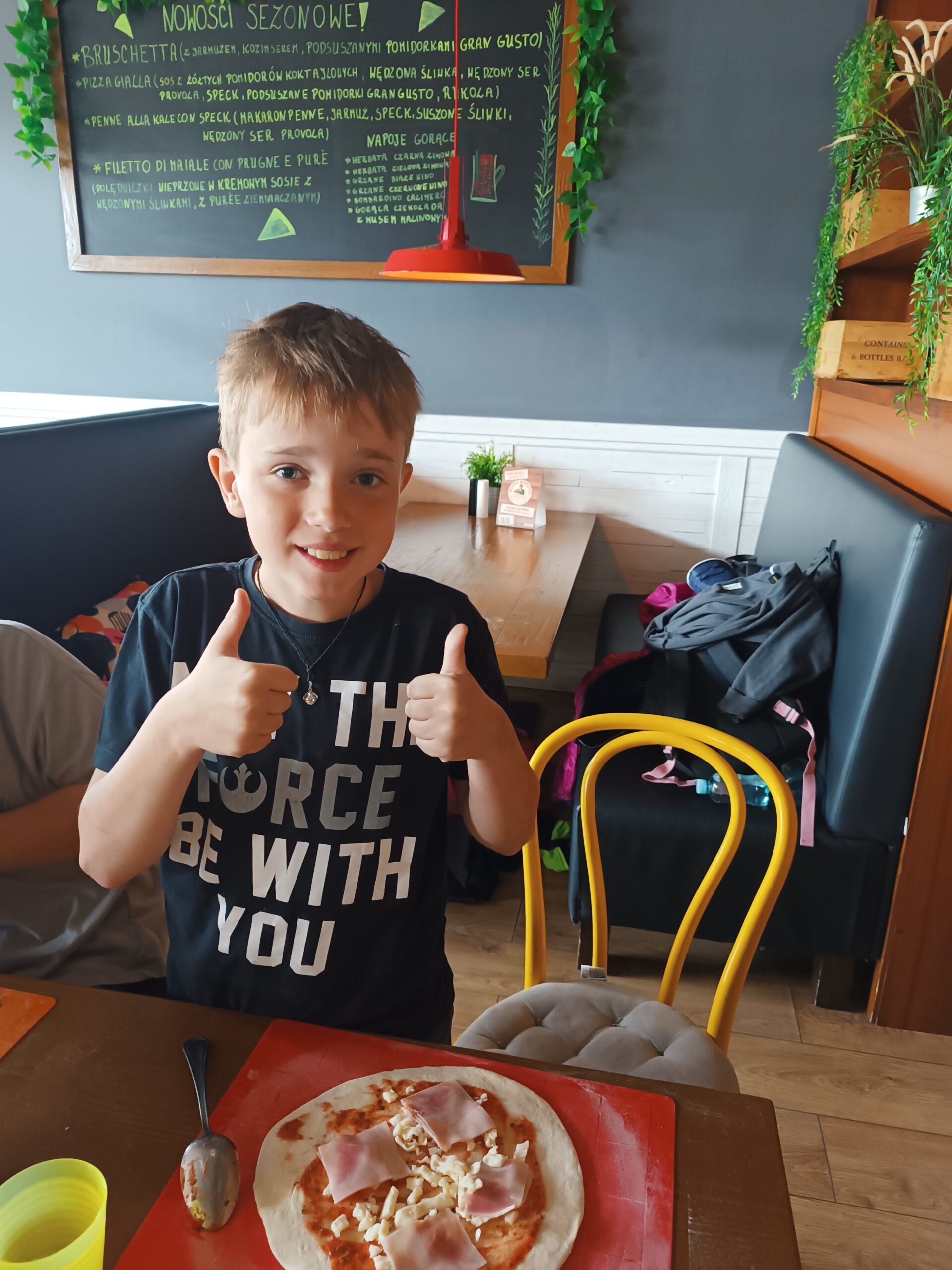 Na zdjęciu widać chłopca z kciukami podniesionymi do góry i z przygotowaną przez niego pizzą